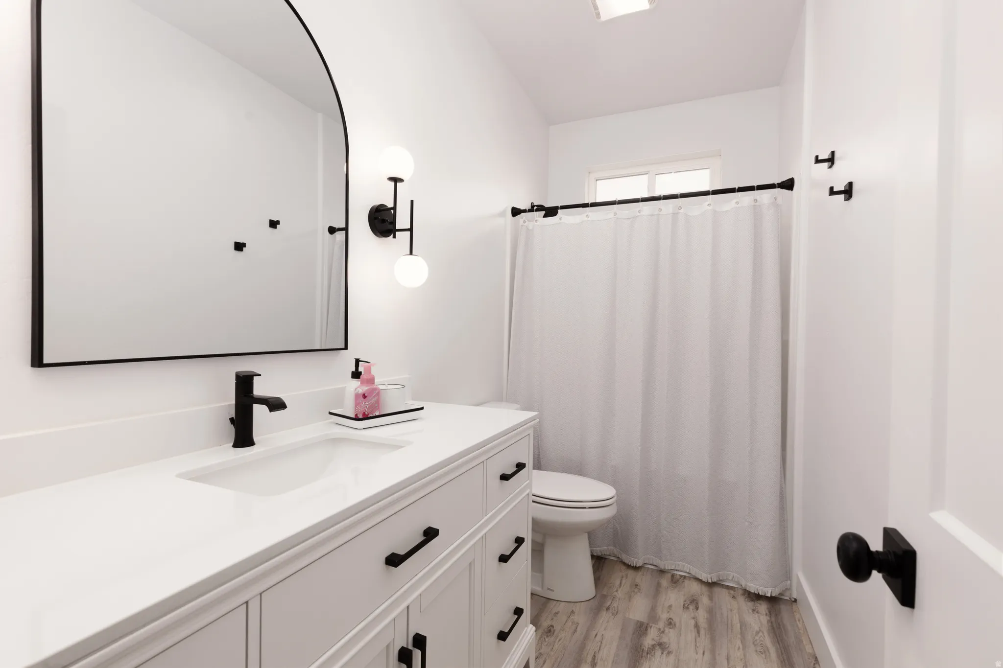 White vanity with integrated sink, matte black faucet, arched mirror, and wood-finish flooring