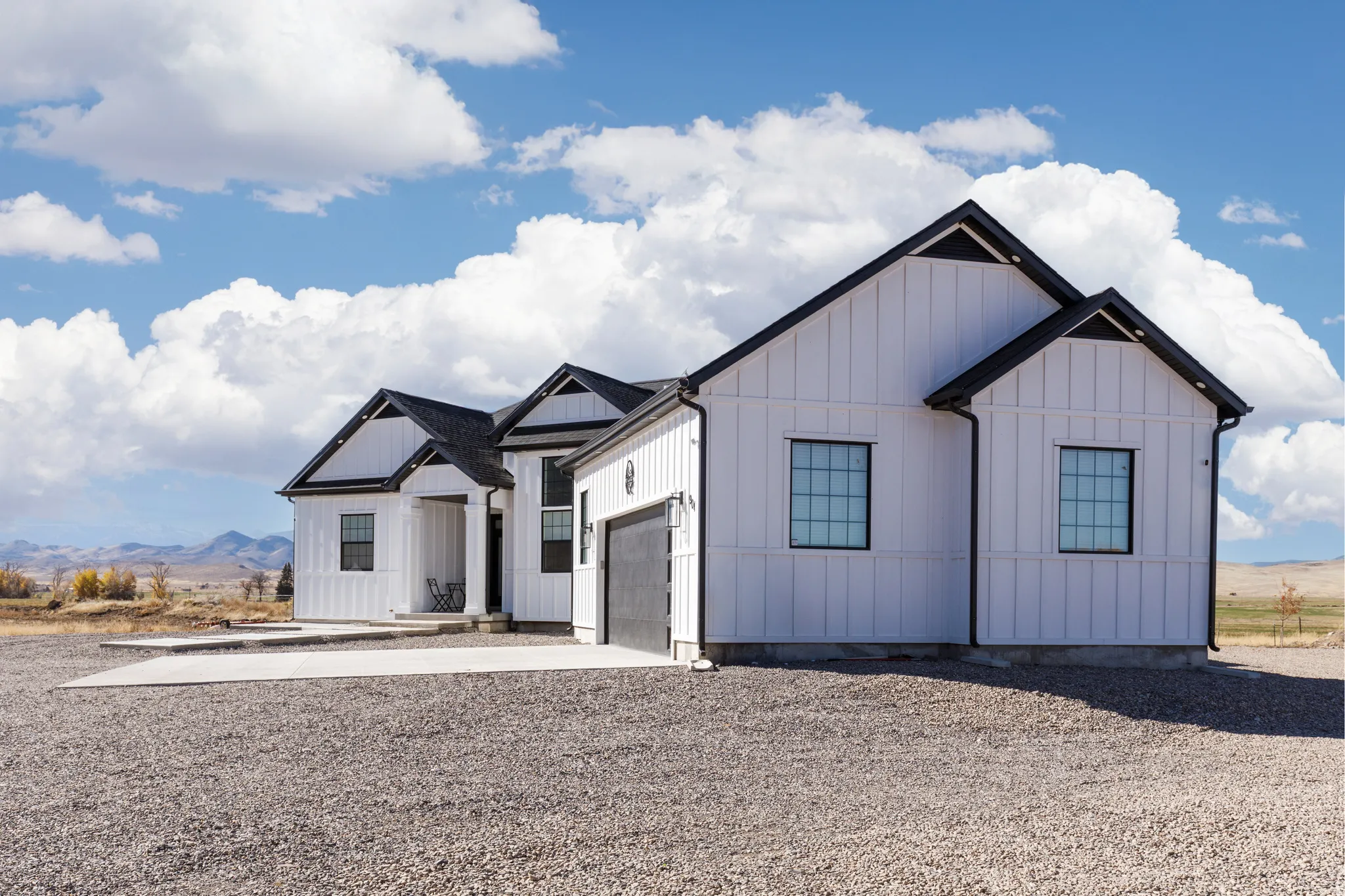 White vertical siding exterior with black trim, multiple gables, attached garage, covered entry, and gravel driveway