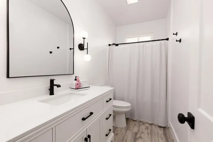 White vanity with integrated sink, matte black faucet, arched mirror, and wood-finish flooring