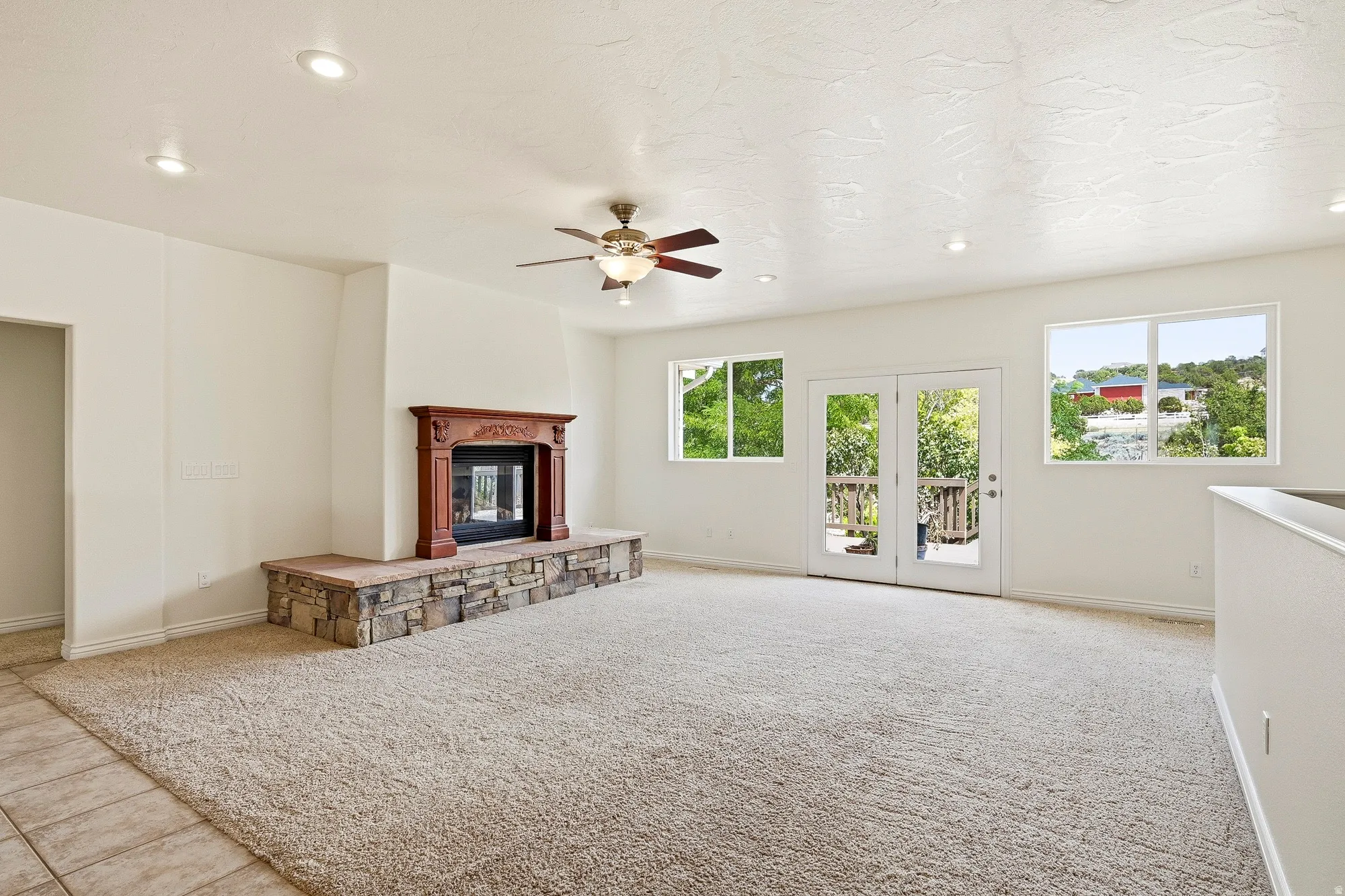 Carpeted living area featuring a fireplace with a stone hearth, a carved wood mantel, a ceiling fan, recessed lighting, and double glass doors opening to a deck