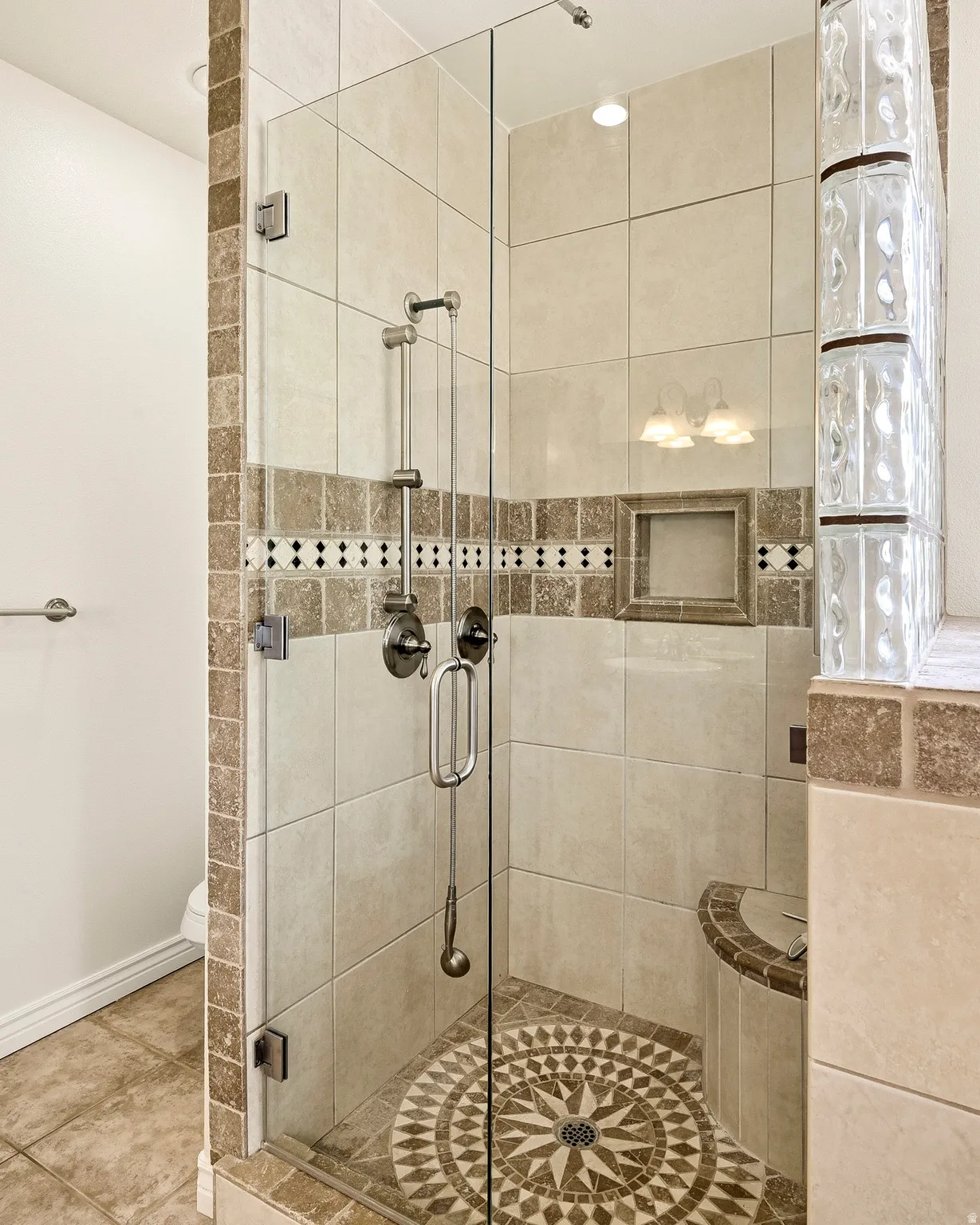 Glass-enclosed shower featuring a tiled floor with a mosaic pattern, a built-in bench, a shower niche, and glass block accent wall