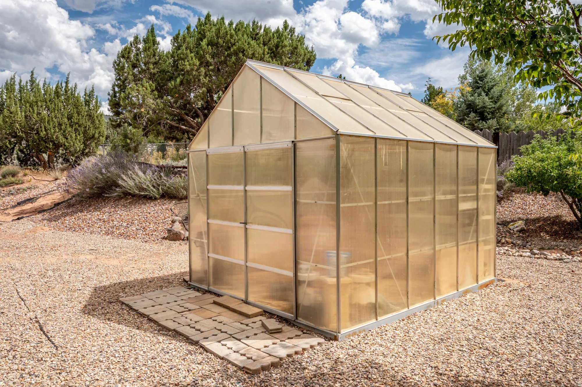 Gable roof greenhouse with translucent panels, set on a gravel-covered yard, with a paver walkway