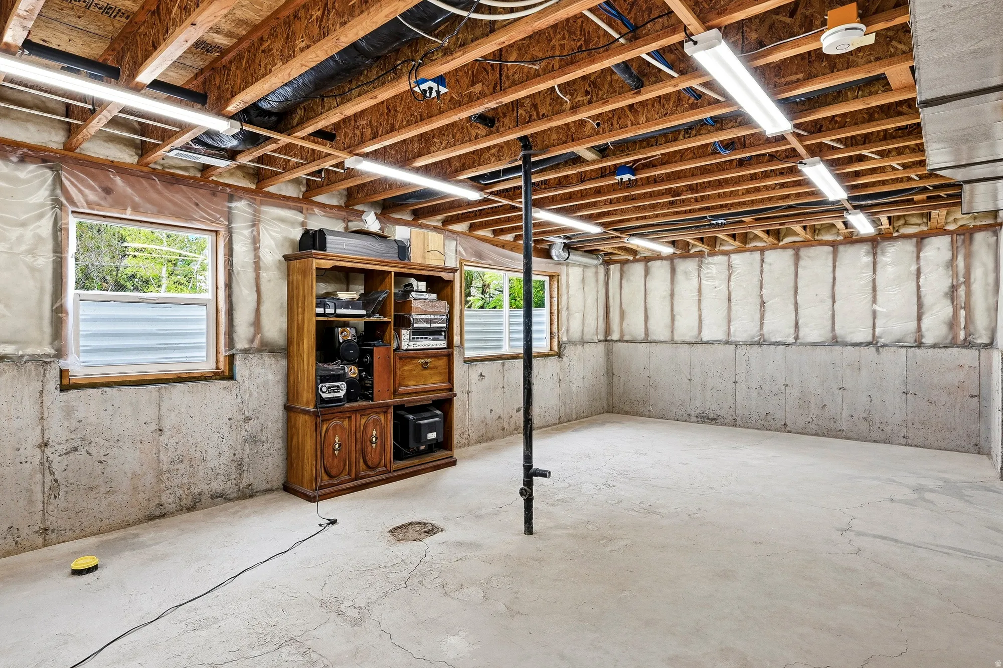 Unfinished basement with exposed wooden joists, concrete floor, multiple windows, and fluorescent lighting