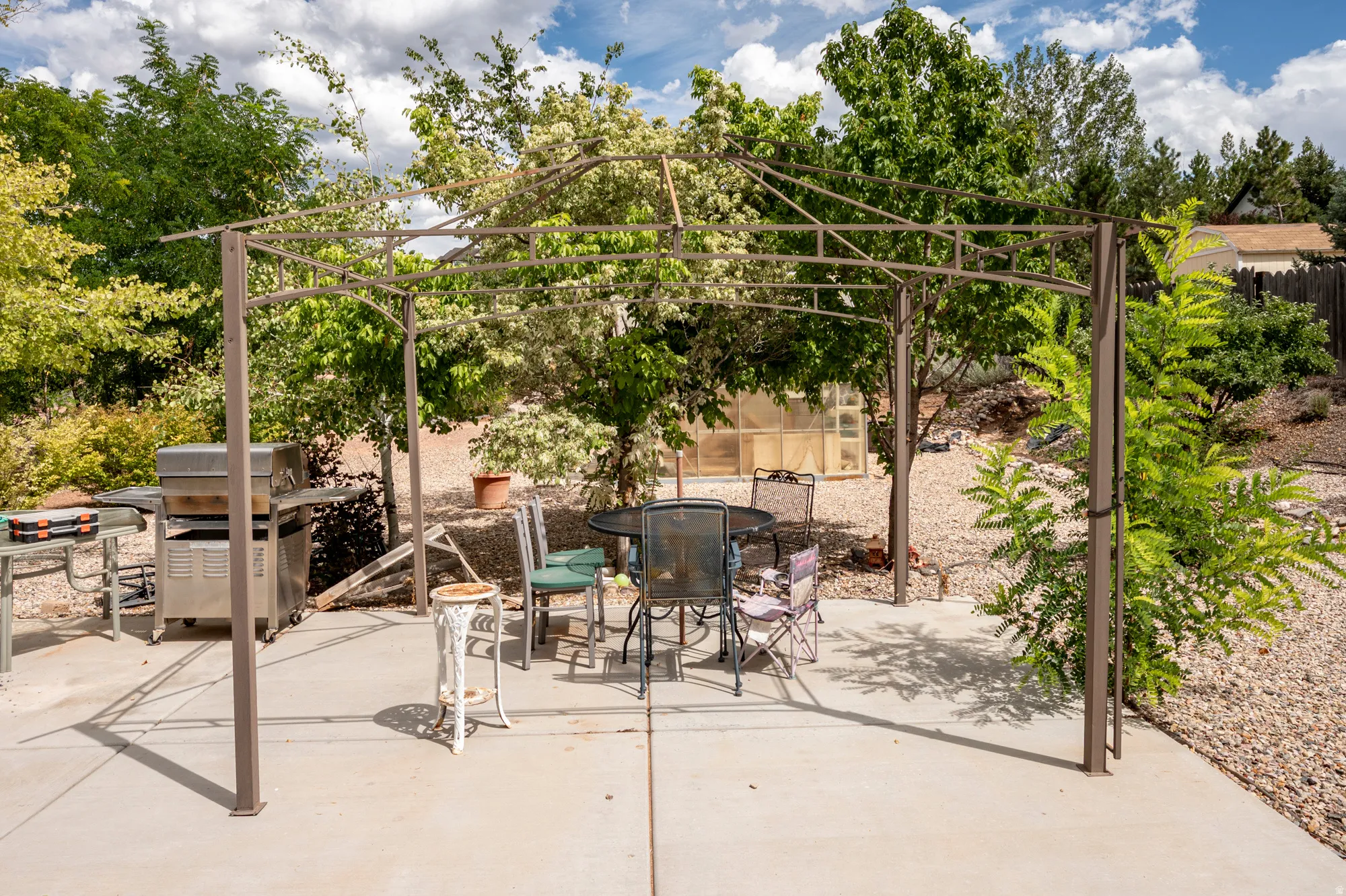 Expansive concrete patio with a permanent metal pergola structure, surrounded by mature trees and a gravel landscape