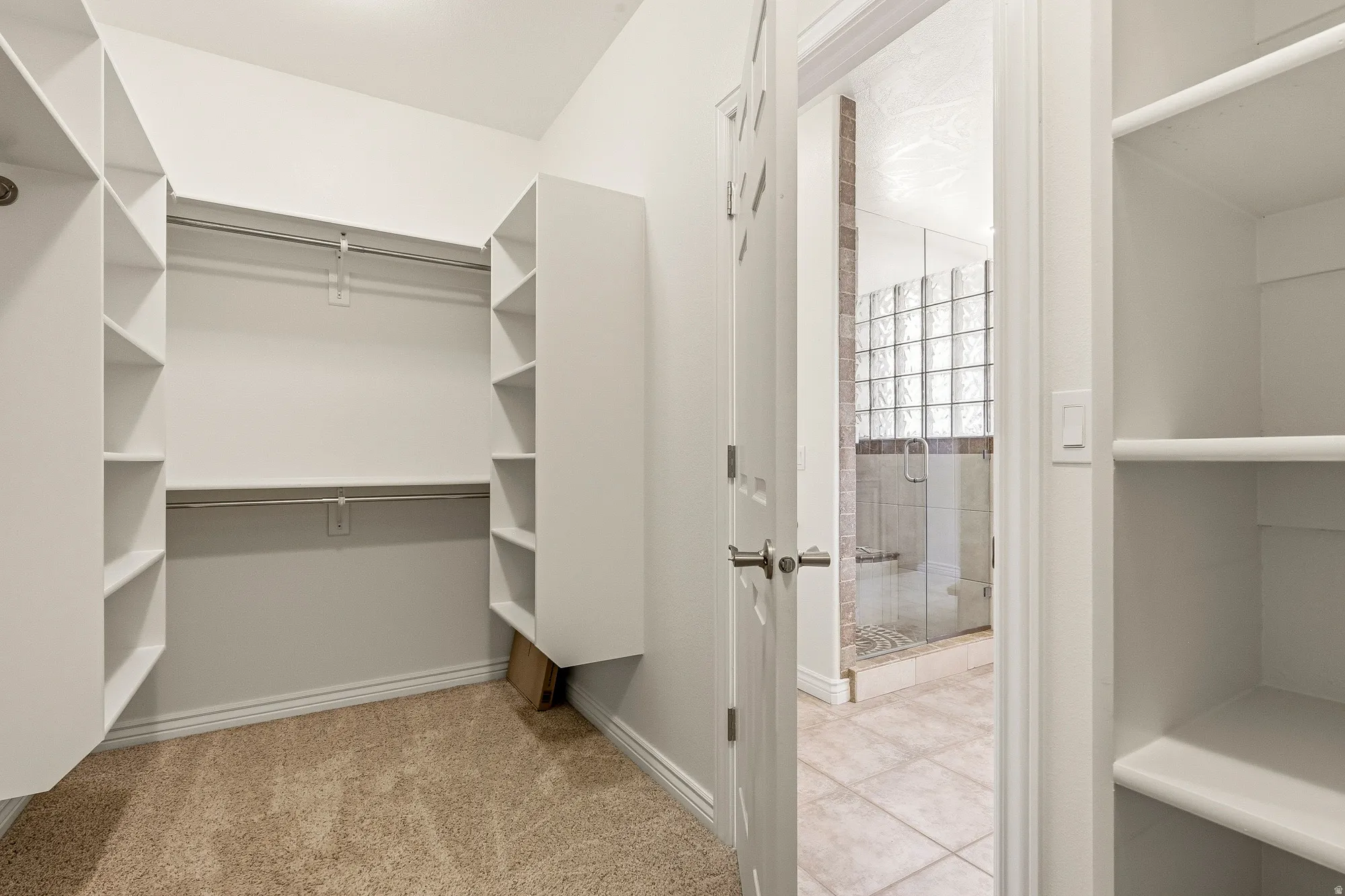 Carpeted walk-in closet with built-in shelving, two hanging rods, and an adjacent bathroom featuring a glass-enclosed shower with tile surround and glass block window