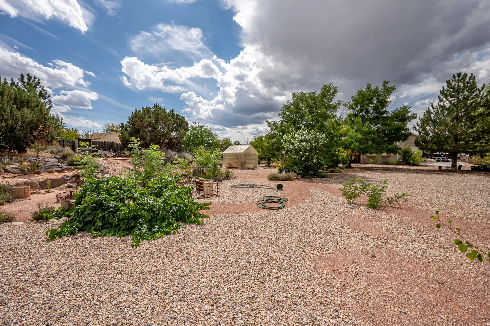 Expansive xeriscaped yard featuring gravel ground cover, mature trees, a dedicated garden area with raised beds, and a greenhouse structure