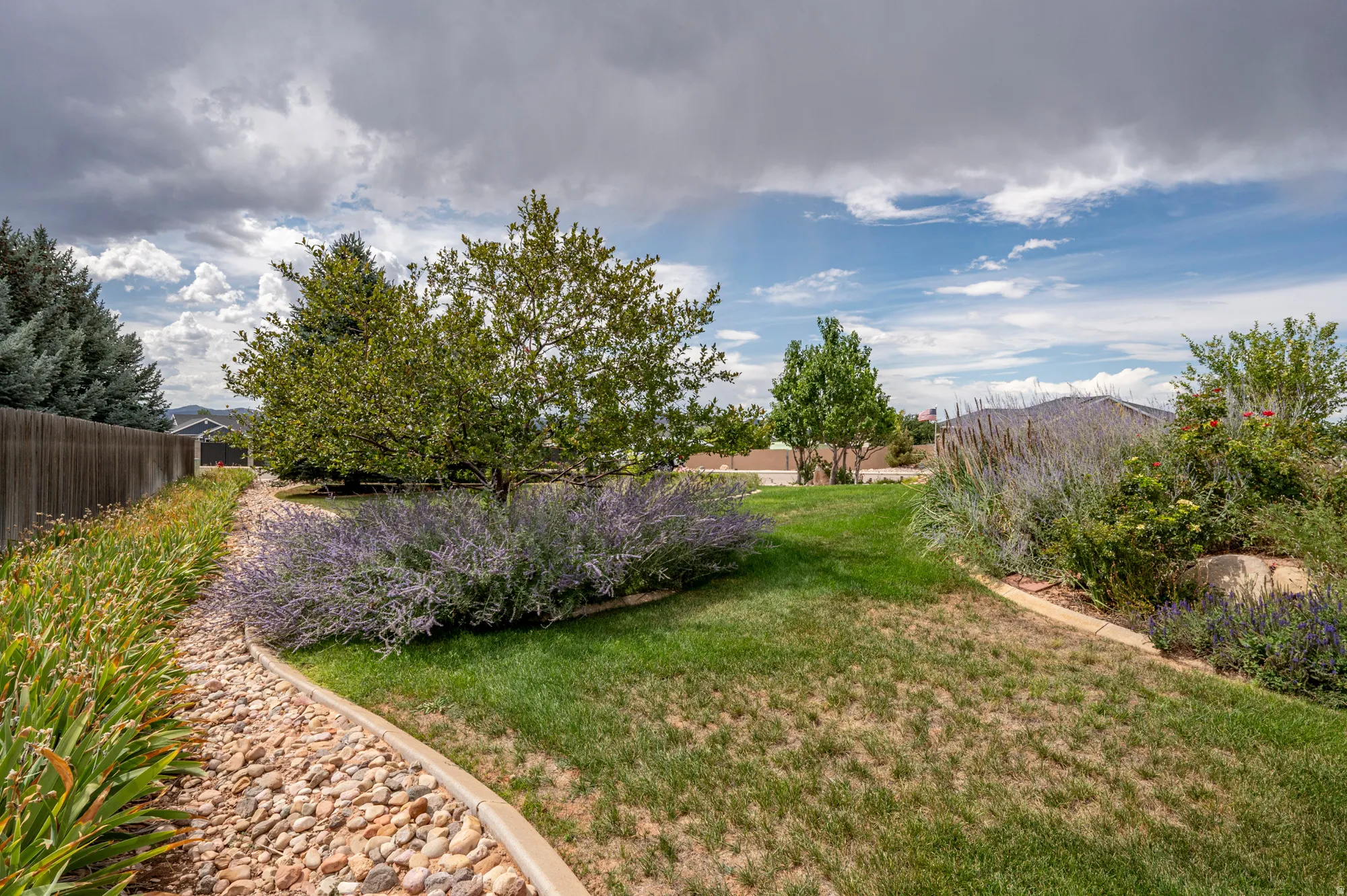 Landscaped backyard featuring a wooden fence, mature trees, and decorative plantings