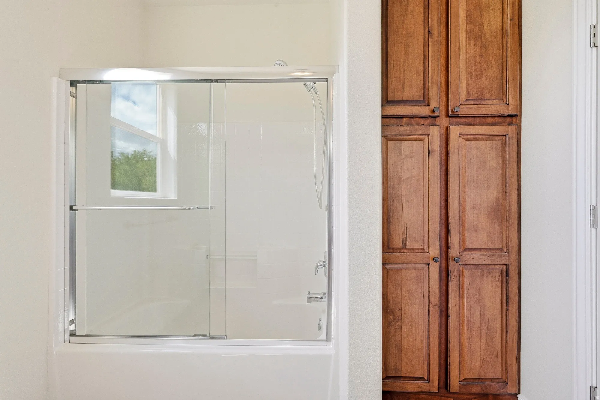 Tub with sliding glass enclosure, integrated shower, and adjacent full-height wood-finish cabinetry