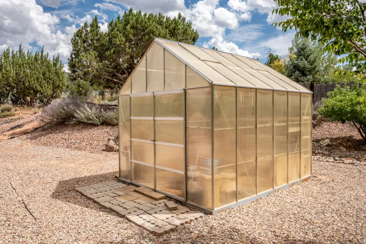 Gable roof greenhouse with translucent panels, set on a gravel-covered yard, with a paver walkway