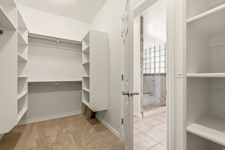 Carpeted walk-in closet with built-in shelving, two hanging rods, and an adjacent bathroom featuring a glass-enclosed shower with tile surround and glass block window