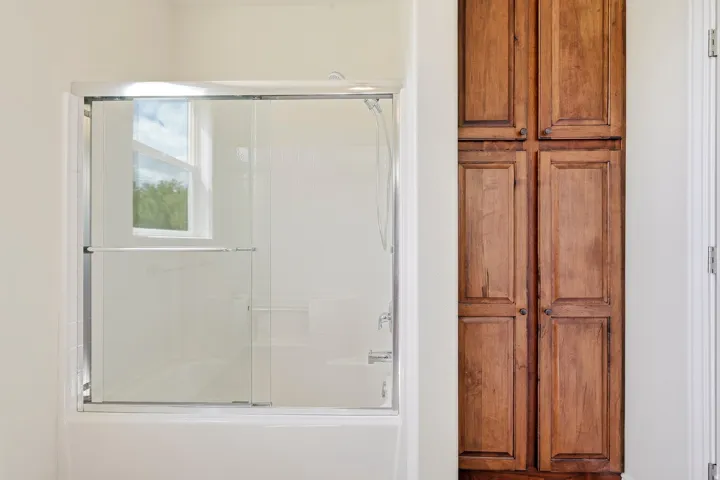 Tub with sliding glass enclosure, integrated shower, and adjacent full-height wood-finish cabinetry