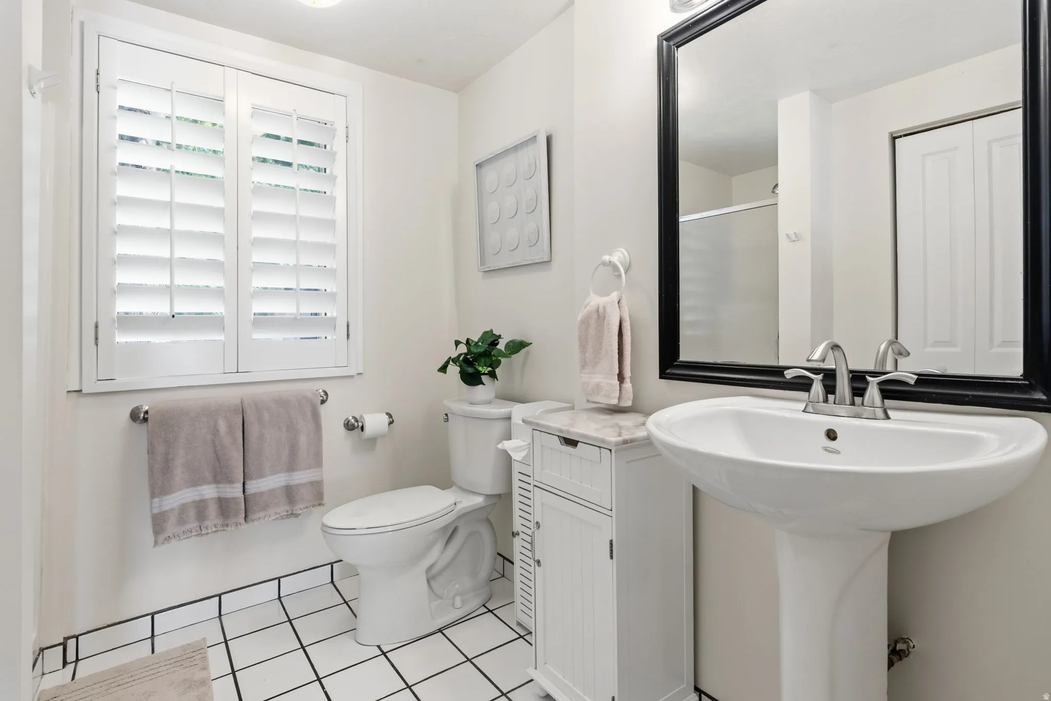 White plantation shutters, pedestal sink with chrome faucet, black-framed mirror, and white tiled flooring