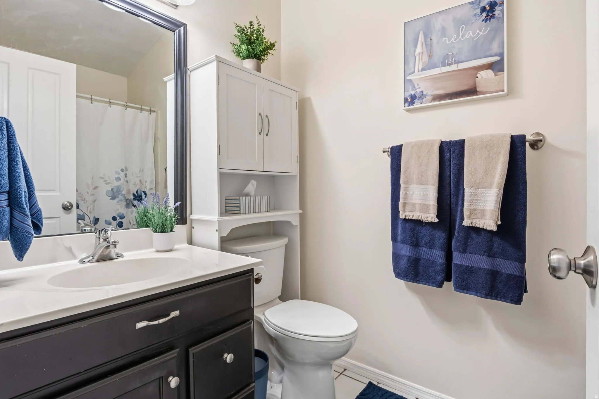 Vanity with a white countertop and undermount sink, dark wood-finish cabinetry, a framed mirror, and a white over-toilet storage cabinet