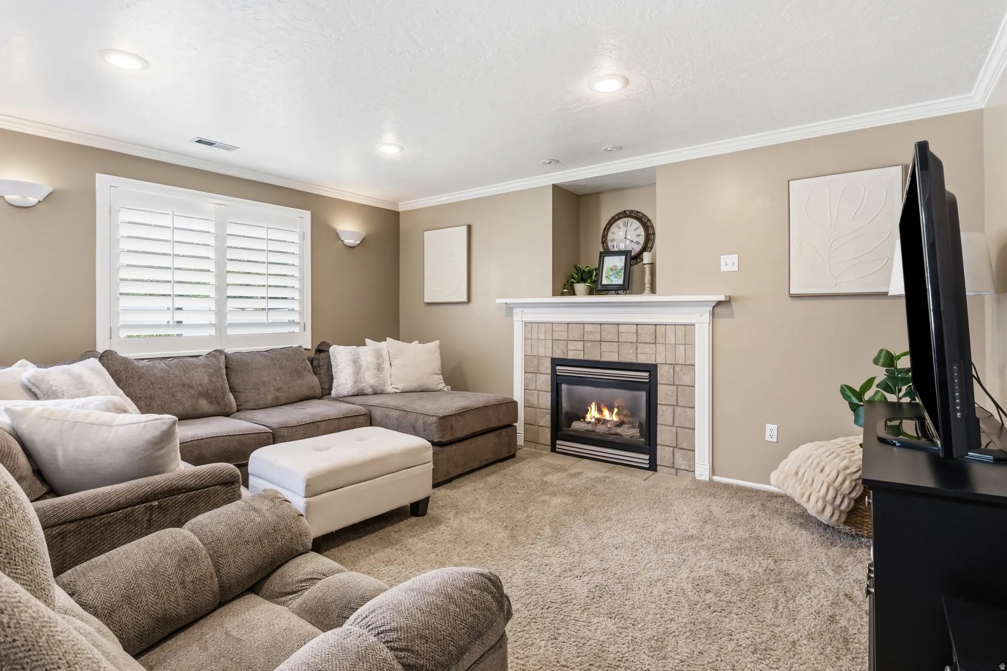 Recessed lighting, crown molding, wall sconces, window with plantation shutters, and a tiled fireplace with a white mantel