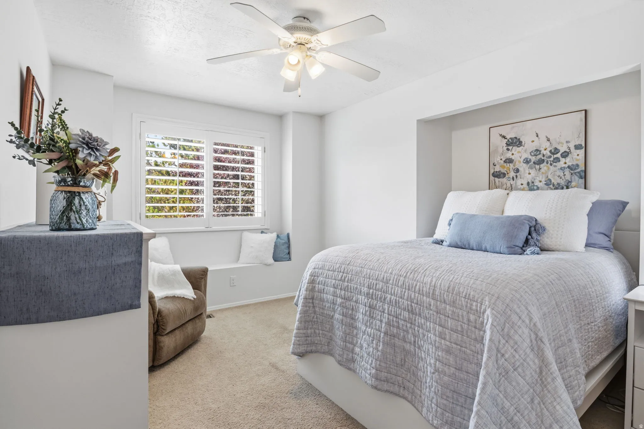 Recessed bed alcove, built-in window bench, white plantation shutters, ceiling fan with integrated lighting, and neutral carpet flooring