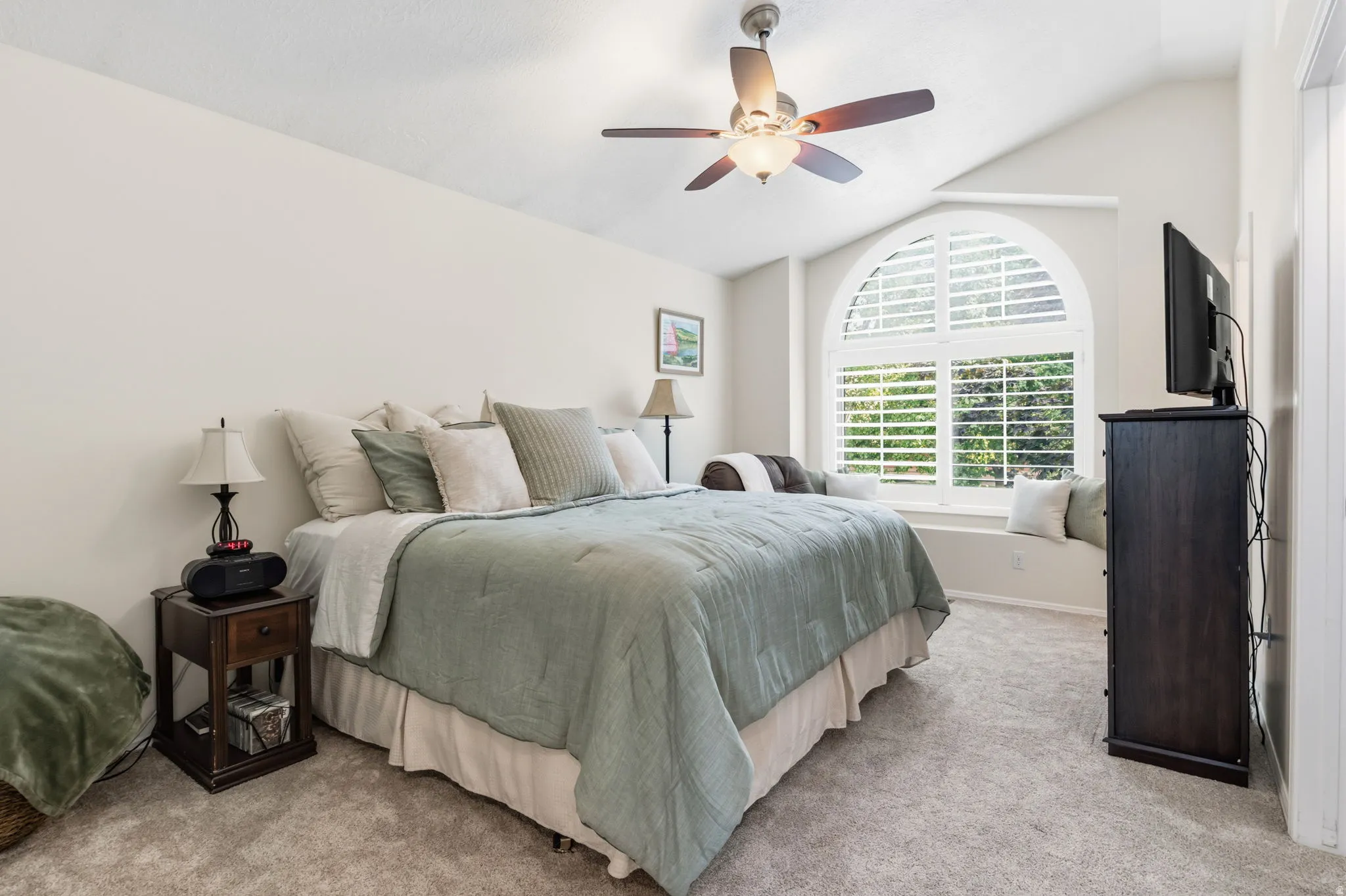 Vaulted ceiling with a ceiling fan, arched window with plantation shutters, window seat, and carpeted flooring