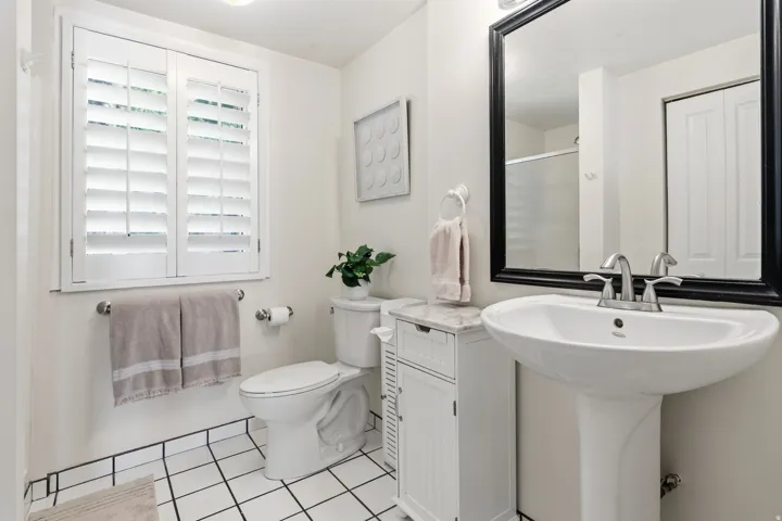 White plantation shutters, pedestal sink with chrome faucet, black-framed mirror, and white tiled flooring