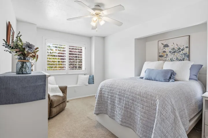Recessed bed alcove, built-in window bench, white plantation shutters, ceiling fan with integrated lighting, and neutral carpet flooring