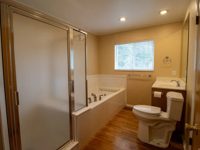 Frosted glass shower enclosure, built-in bathtub, vanity with integrated sink, wood-finish flooring, and recessed ceiling lighting