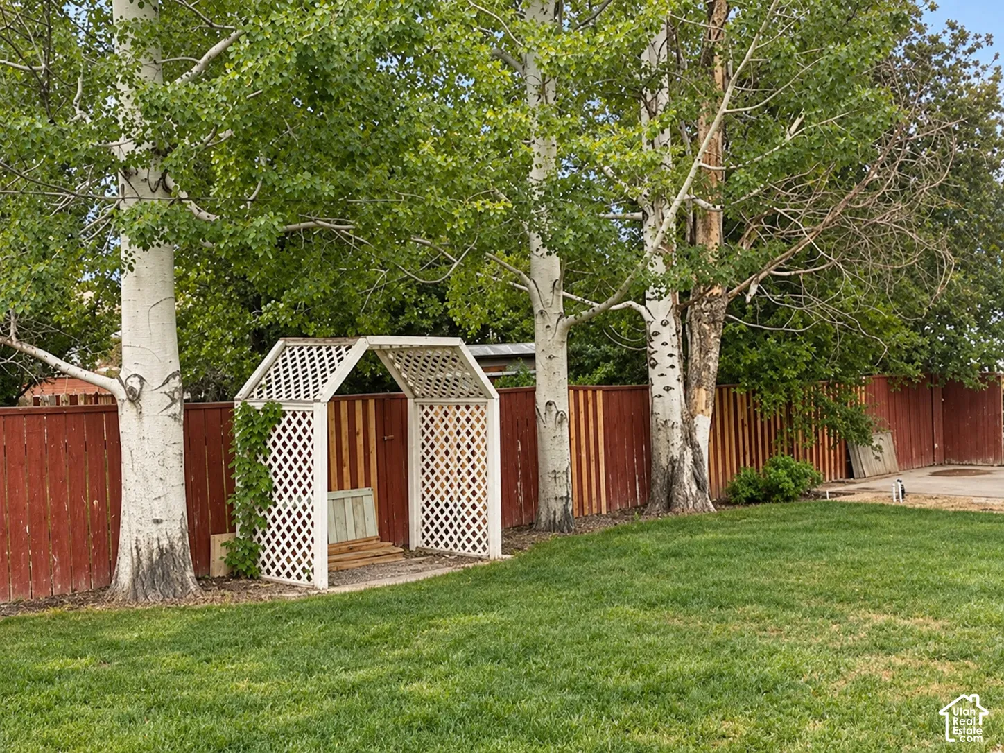 Backyard featuring a manicured lawn, white wood lattice arbor, mature trees, and a full-height red wood privacy fence