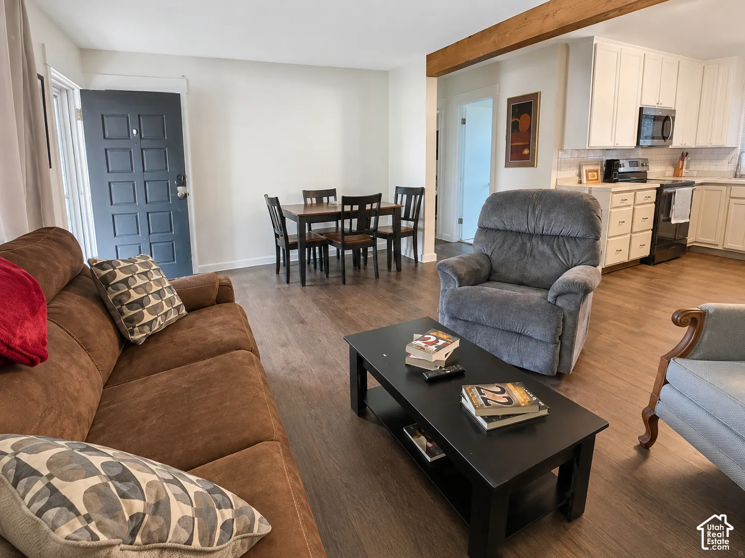Open-concept interior featuring wood-finish flooring, exposed structural ceiling beam, white shaker-style cabinetry, and a paneled entry door