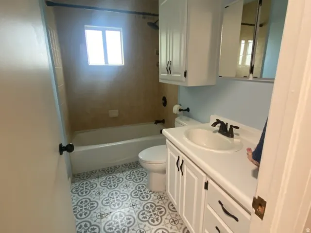 Patterned floor tile, white vanity with black hardware, integrated sink, overhead wall cabinetry, and a bathtub with tiled surround