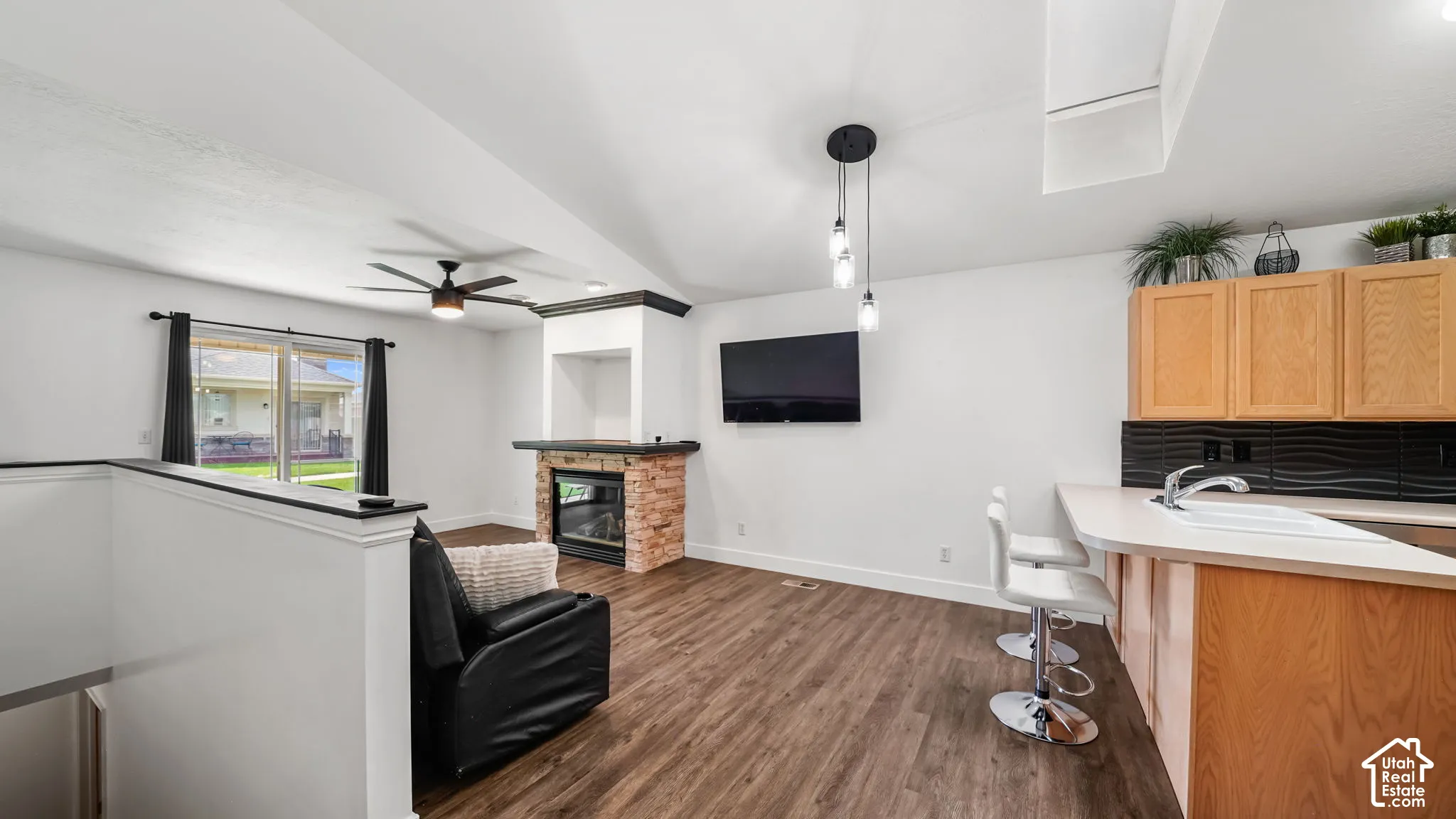 Wood-finish flooring throughout, featuring a fireplace with stone surround, a ceiling fan, and a kitchen area with upper cabinetry and a sink