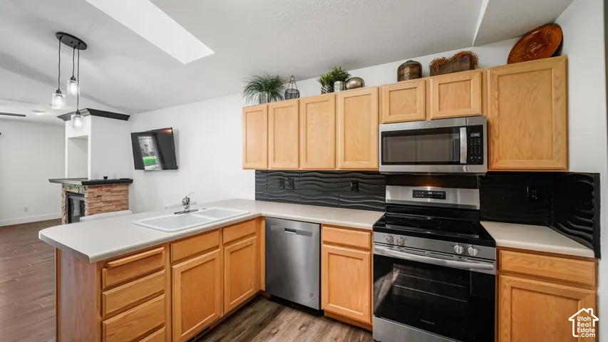 Open kitchen with light wood cabinetry, stainless steel appliances, a double basin sink, and a textured black backsplash