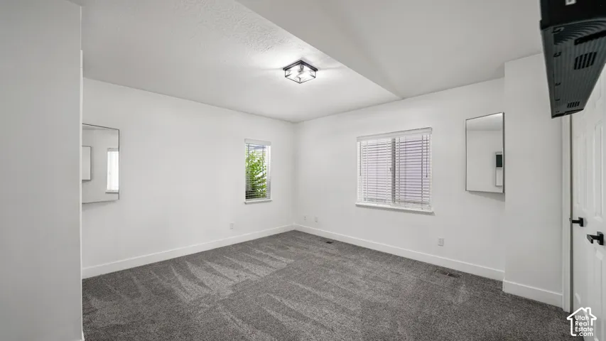 Neutral wall paint, textured ceiling, two windows with blinds, gray patterned carpeting, and a flush-mount ceiling light