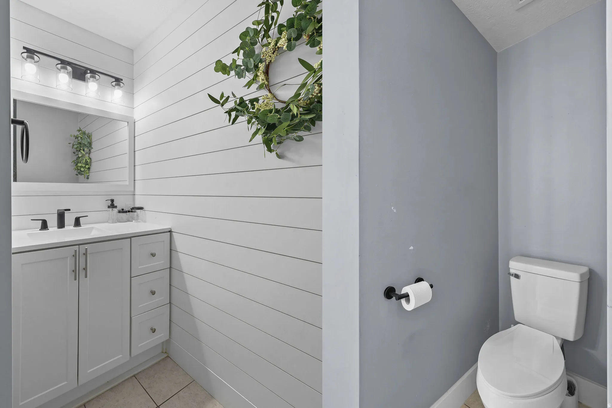White shiplap accent wall, vanity with white countertop, chrome hardware, rectangular mirror, and neutral tile flooring