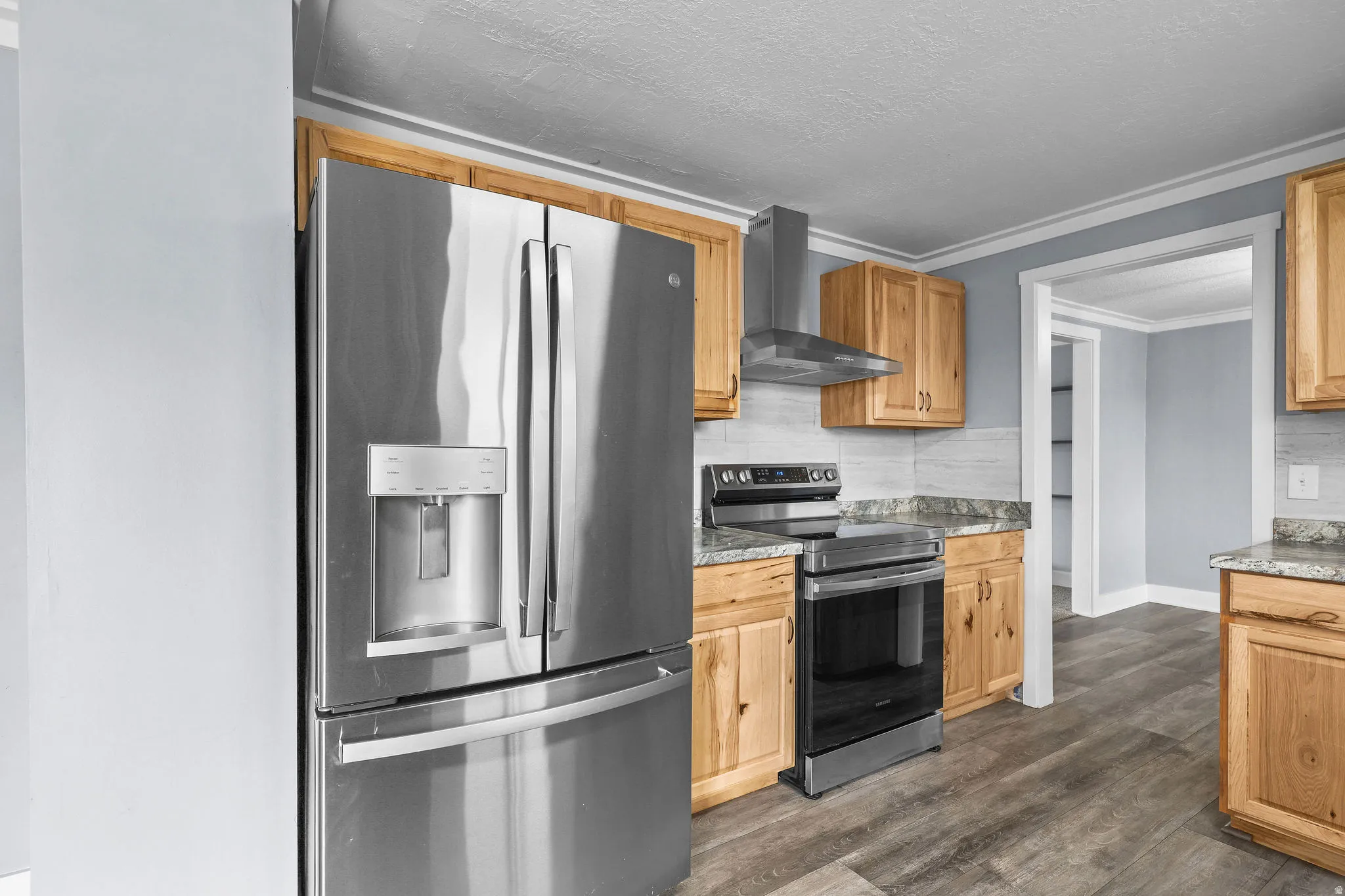 Stainless steel refrigerator, stainless steel range with overhead vent hood, light wood-finish cabinetry, patterned stone-finish countertops, light tile backsplash, and wood-finish flooring