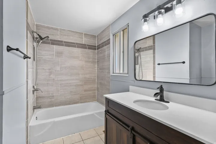 White bathtub with tile surround, dark wood-finish vanity with white countertop, and rectangular mirror