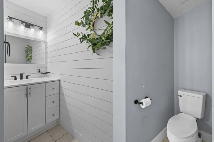White shiplap accent wall, vanity with white countertop, chrome hardware, rectangular mirror, and neutral tile flooring