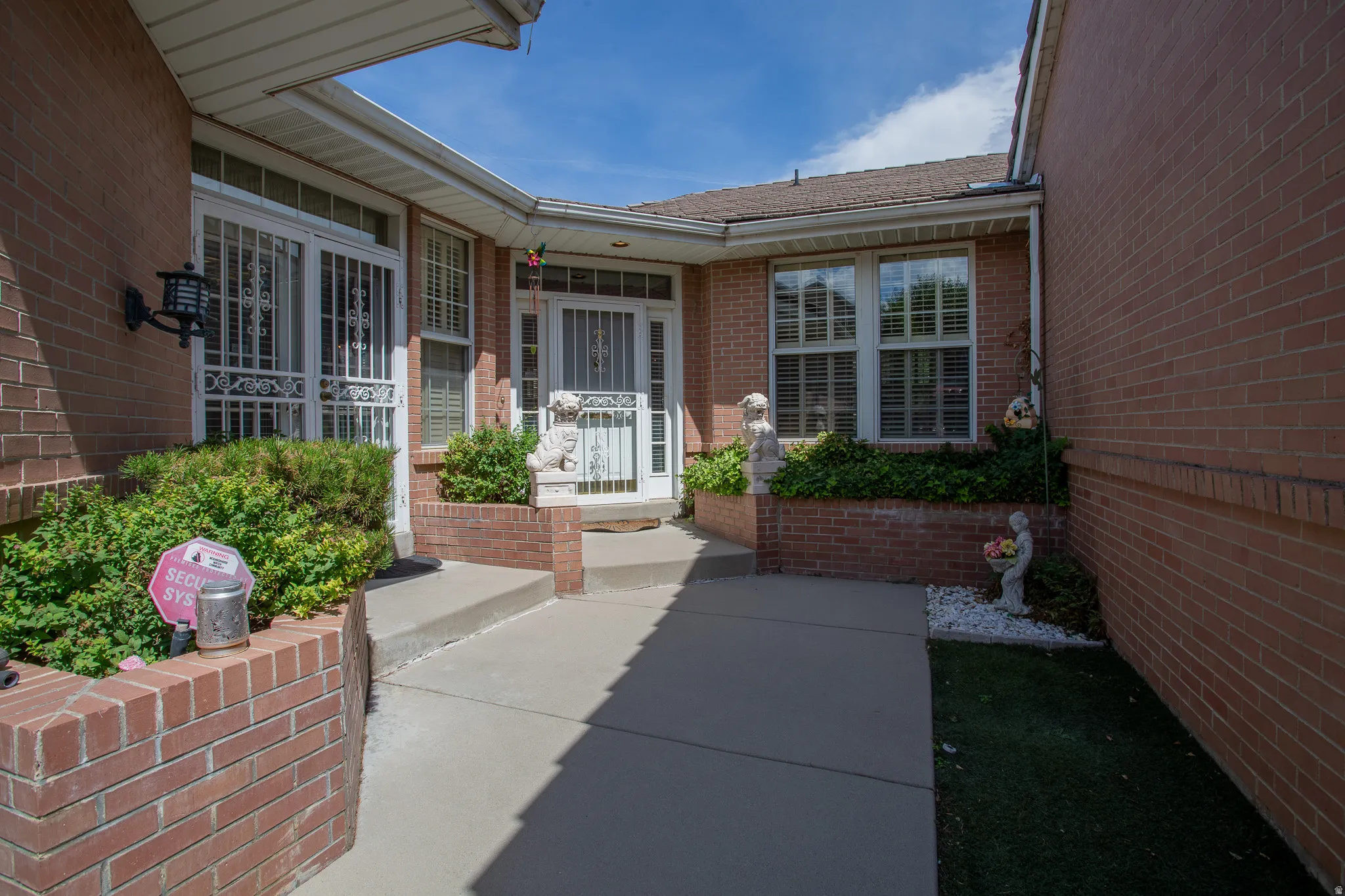 Brick exterior with concrete walkway, mature shrubbery, and window shutters