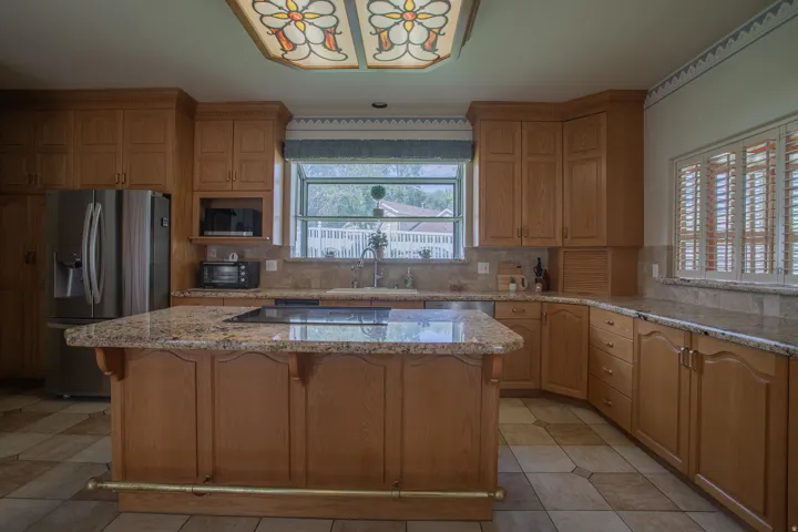 Expansive kitchen featuring an island with cooktop, granite countertops, wood cabinetry, stainless steel refrigerator, and tiled flooring
