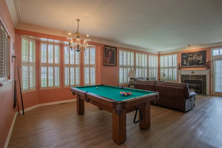 Spacious room featuring wood-finish flooring, a bay window with plantation shutters, an ornate chandelier, and a fireplace with a stone surround