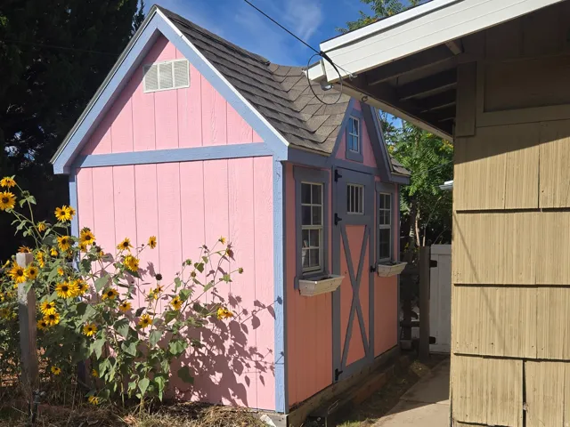 Pink and blue painted shed with solar electricity, featuring a gabled roof, two windows, and dark-shingle roofing.