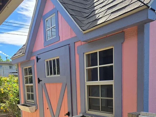 Gable-roofed shed with shingle roofing, vertical plank siding, multiple windows, and decorative window boxes, has solar electricity.