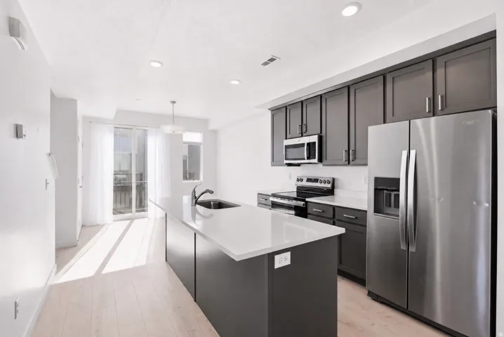 Modern kitchen featuring a large island with an undermount sink, stainless steel appliances, dark wood-finish cabinetry, and light-tone flooring
