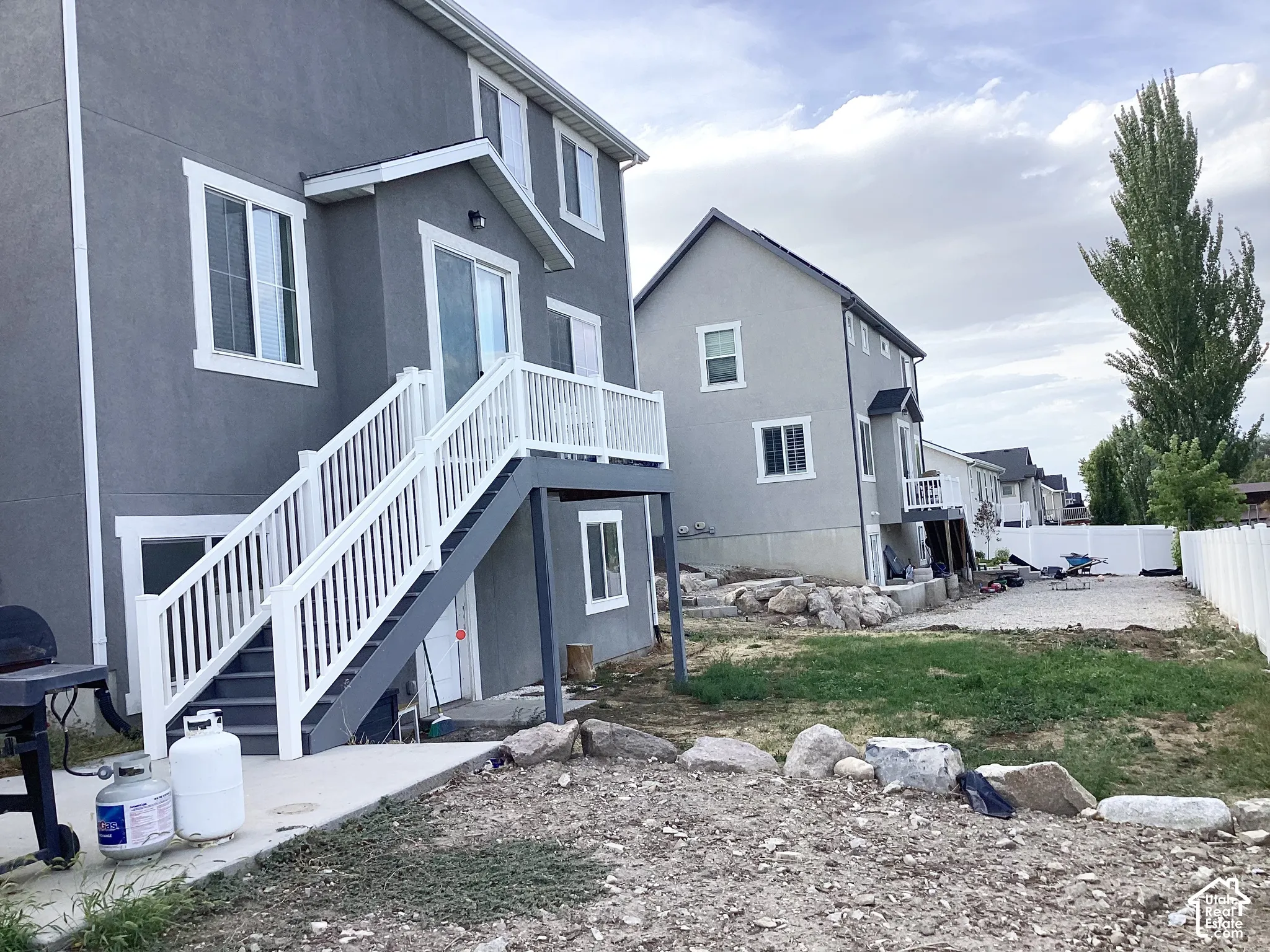 Gray stucco exterior with white trim, upper-level balcony, white railing system, lower-level concrete patio, and gravel landscaping