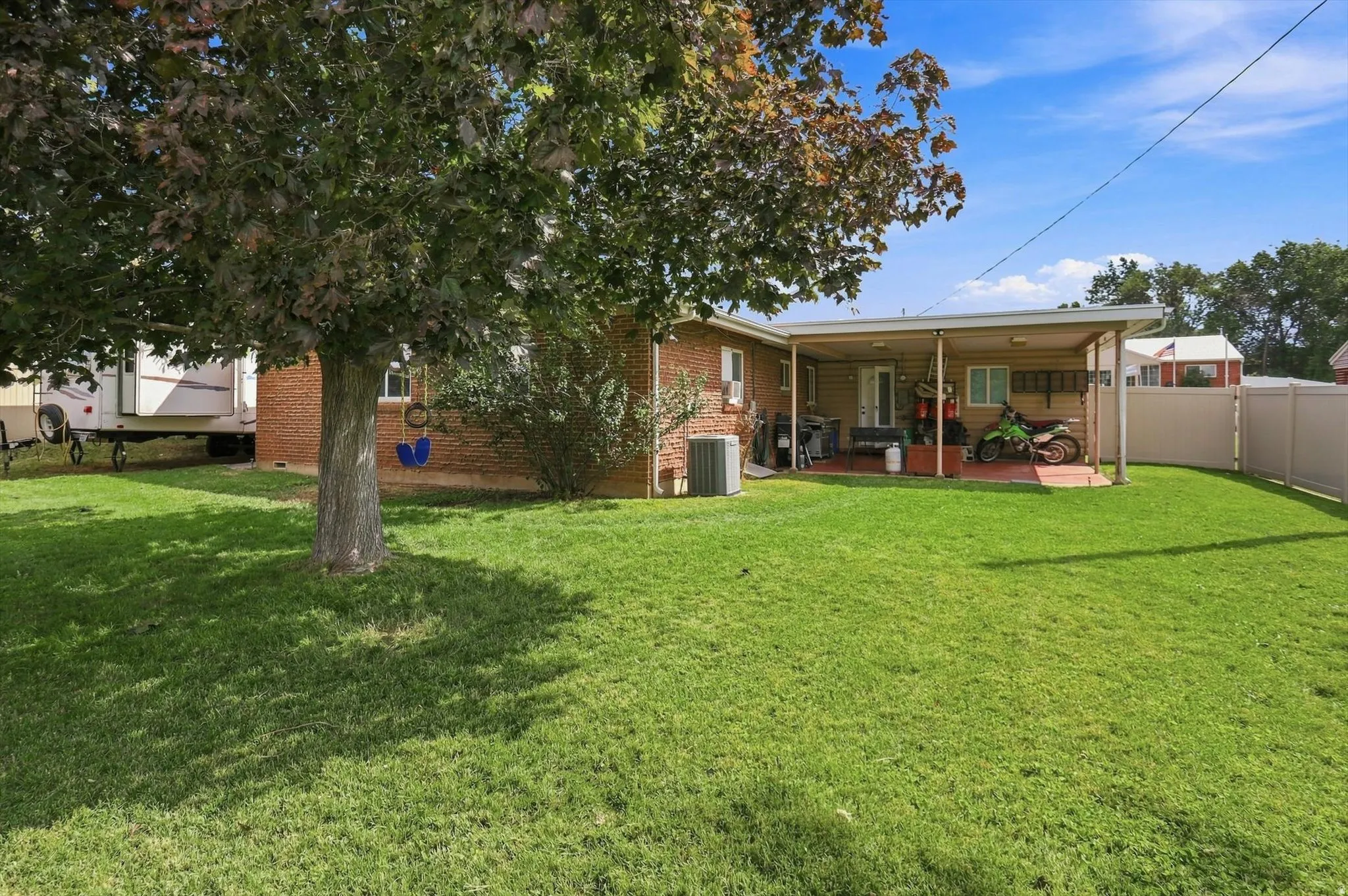 Expansive grassy lawn with a mature tree, brick exterior, covered patio, and privacy fencing