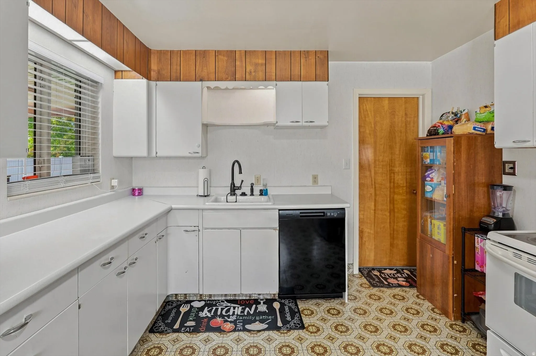 White cabinetry, new black dishwasher, wood-finish door, patterned floor tile, and a window with blinds