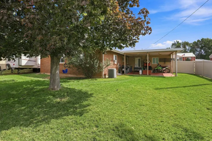 Expansive grassy lawn with a mature tree, brick exterior, covered patio, and privacy fencing