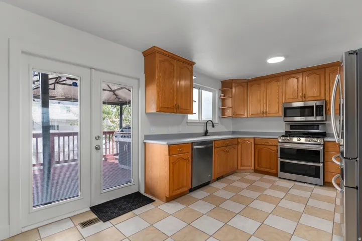 Spacious kitchen featuring warm wood cabinetry, stainless steel appliances including double wall ovens, a large center island with sink, ceramic tile flooring, and glass-panel doors opening to an outdoor deck