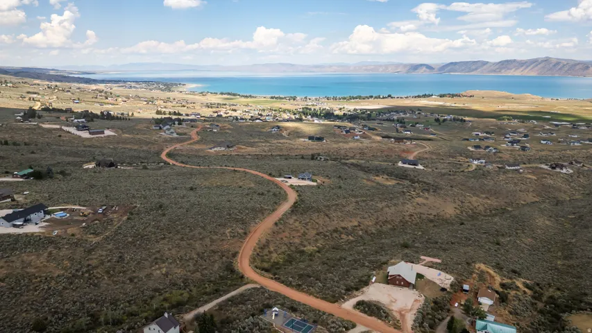 Expansive landscape featuring an unpaved road, scattered residential structures, and distant water body with mountains
