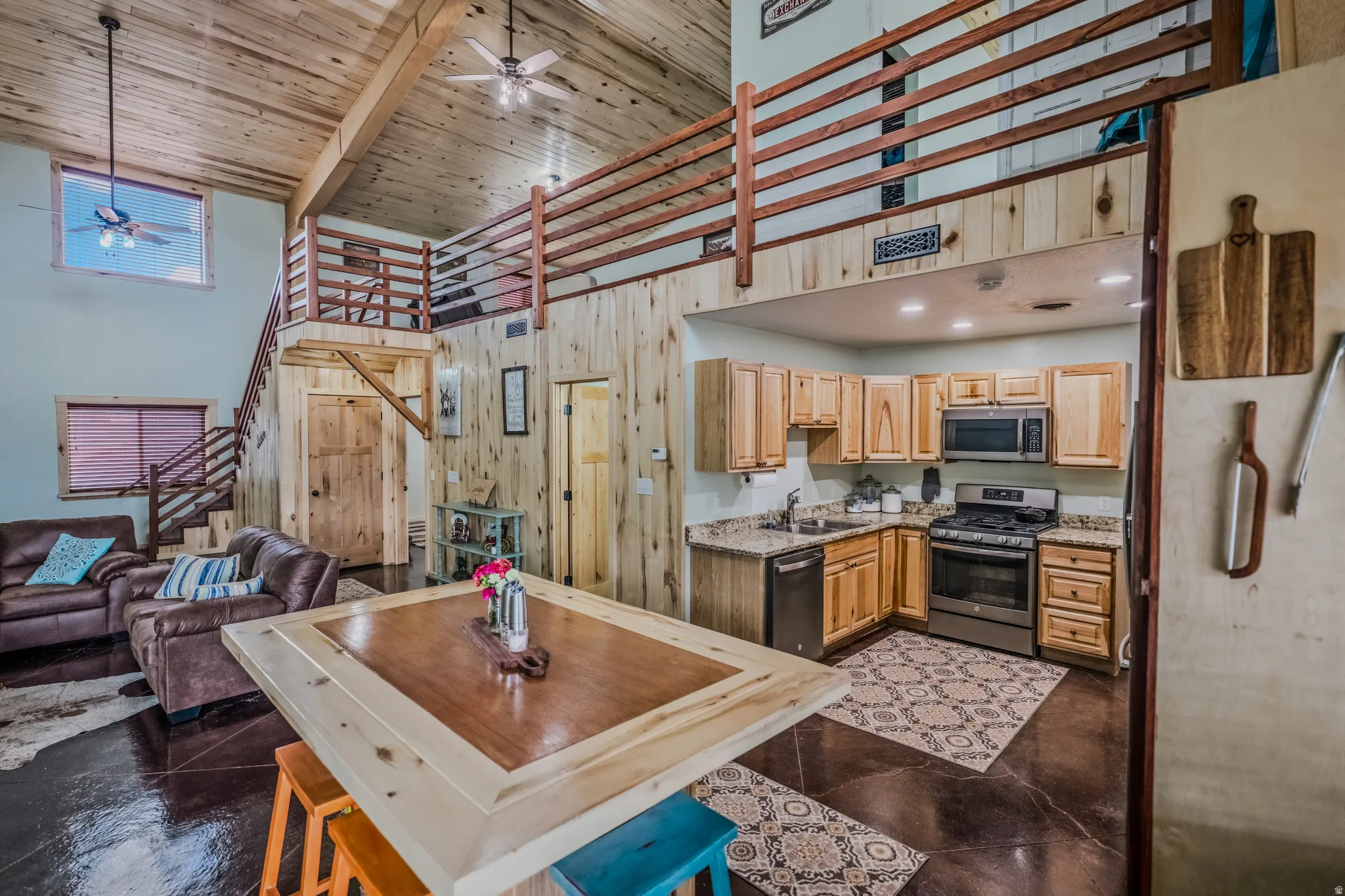Open concept living area with vaulted wood-paneled ceilings, wood-finish flooring, and a kitchen featuring wood cabinetry, stainless steel appliances, and stone-finish countertops