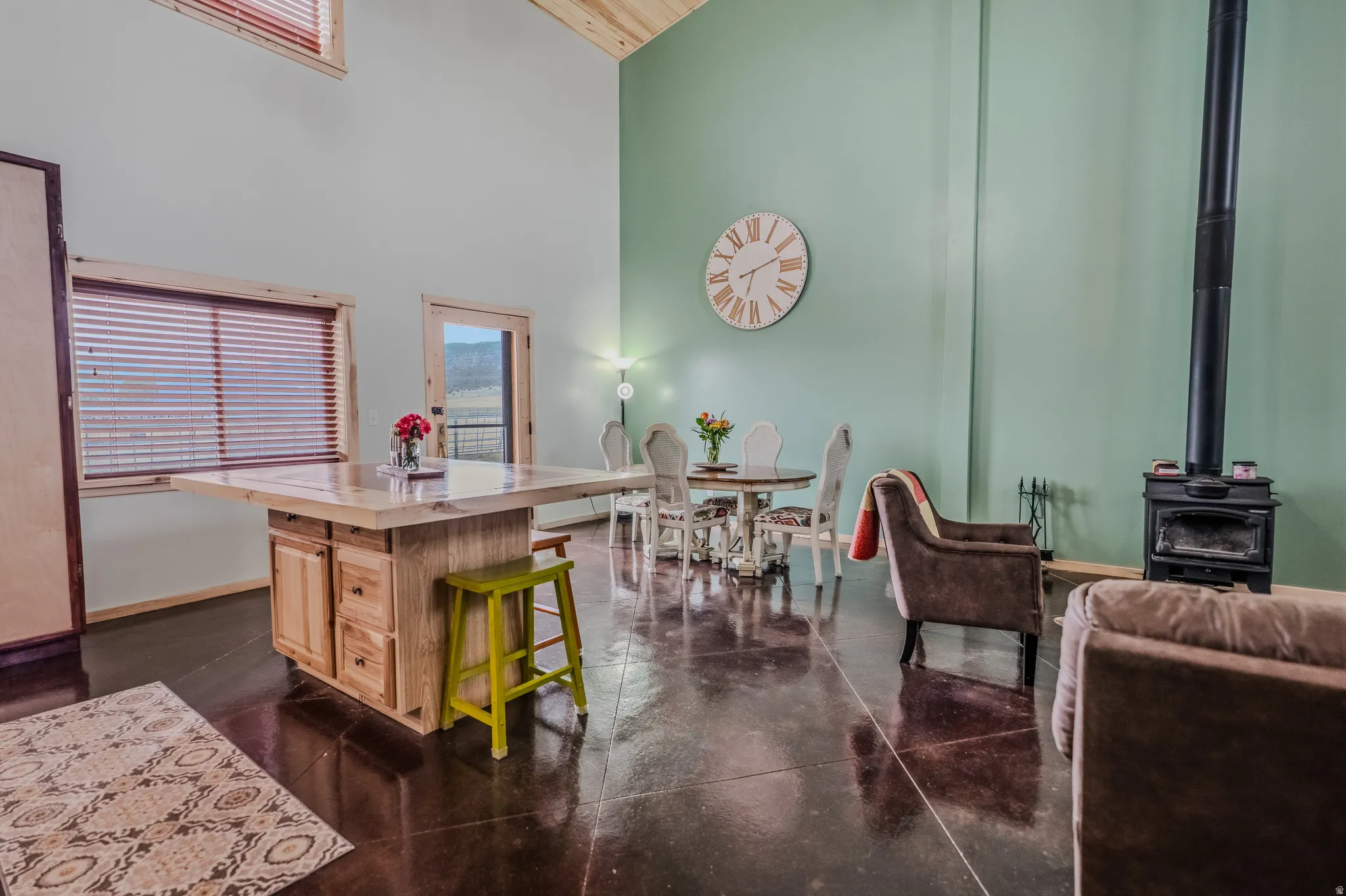 Spacious room with high ceilings, polished concrete flooring, exposed wood beams, a wood-burning stove, and a wood-framed window with blinds