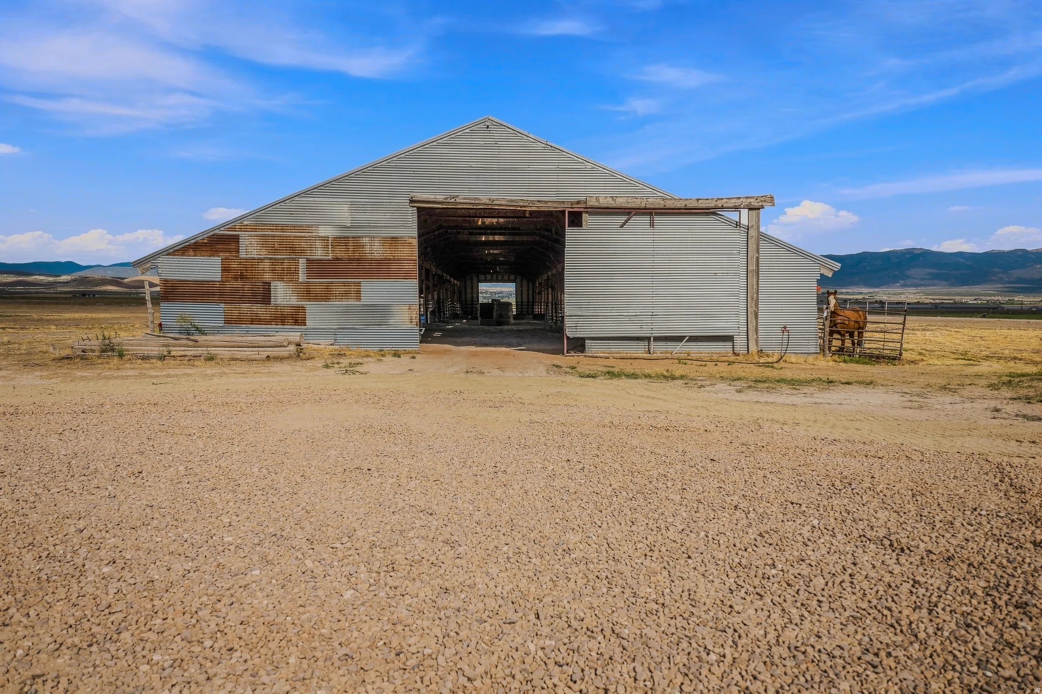Corrugated metal barn with an open central bay, a gravel foreground, and a distant mountainous horizon