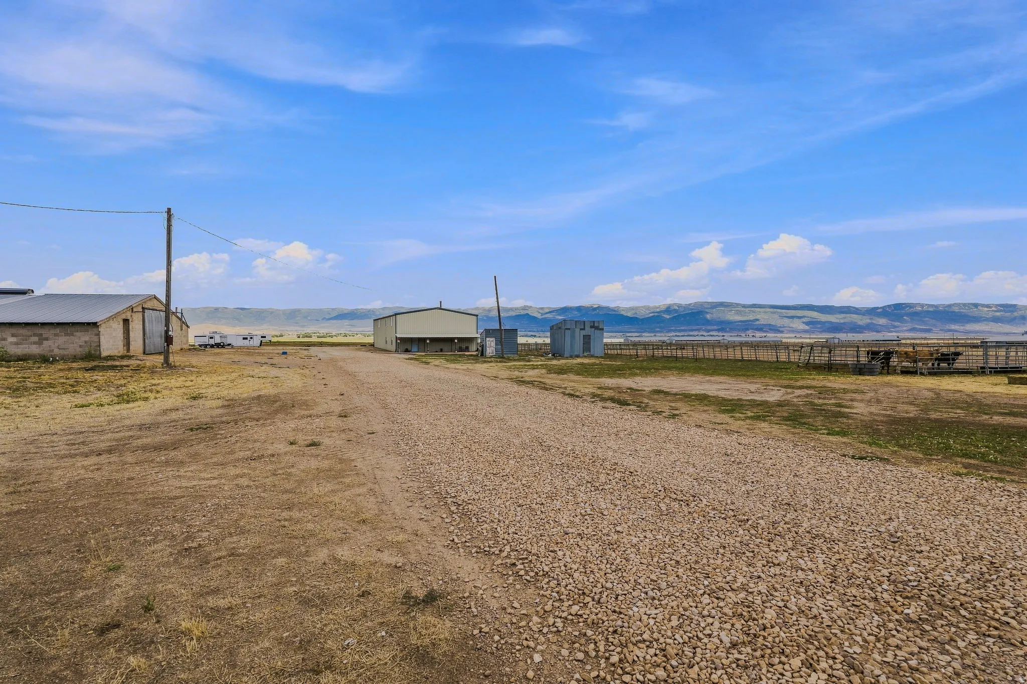 Gravel driveway leading to two large outbuildings, fenced areas, and distant mountainous terrain