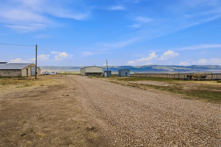 Gravel driveway leading to two large outbuildings, fenced areas, and distant mountainous terrain